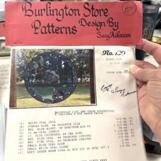 Red storefront pattern book cover reading 'Burlington Store Patterns' with 'Design By Suzy Aikinman' and a circular stained-glass window pattern image shown beside a hand holding the booklet.