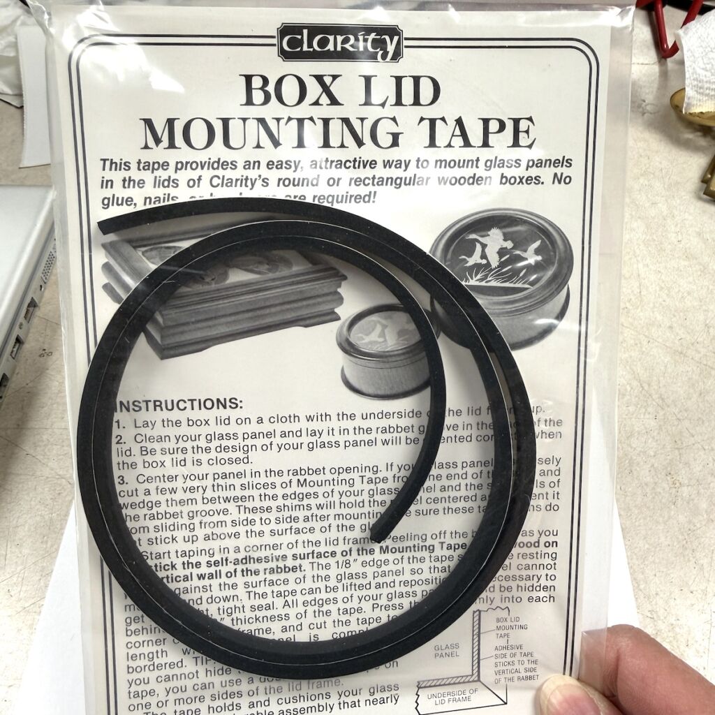 Clarity Box Lid Mounting Tape packaging with two black circular rubber strips laid over instruction sheet for mounting glass panels in box lids.
